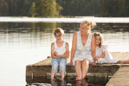 Beautiful young mother and her two kids sitting on a jetty in the summer sun, bathe the feet in the water, and enjoy a beautiful day in midsummer.の写真素材