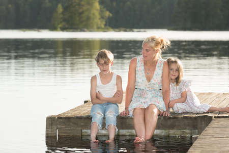 Beautiful young mother and her two kids sitting on a jetty in the summer sun, bathe the feet in the water, and enjoy a beautiful day in midsummer.の写真素材