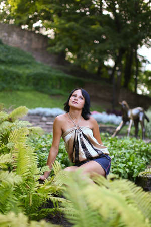 Portrait of young sadness woman which is sitting near to bush of fern in central park against...