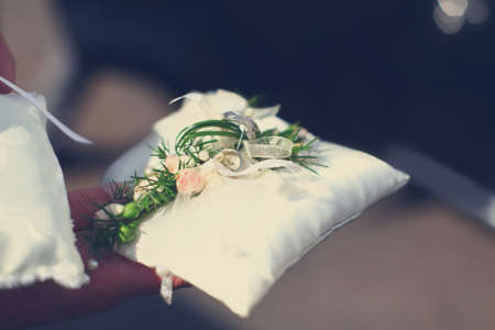 Wedding golden rings are holding in hand on white soft pillow from silk decorated with flowers.の写真素材