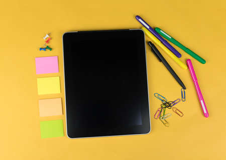 Top view of a tablet and school supplies like colored markers, sticker and clipers on yellow...