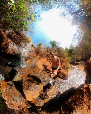 Man in glasses and swim trunks at the waterfall with 360 camera on a sunny day