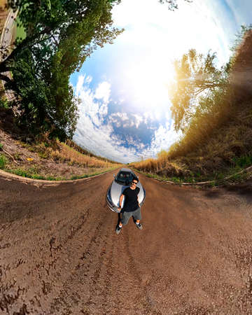 Man black next to a car in countryside mini planet. 360 camera footage with black man, trees, white clouds and soft blue sky.の写真素材