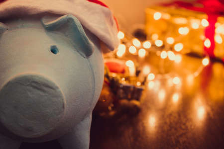 Piggy bank with Santa Claus hat on blurred christmas background with ornaments, gifts and light. Space for text.の写真素材