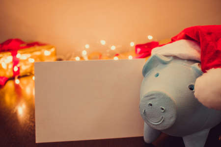 Piggy bank with Santa Claus hat on blurred christmas background with ornaments, gifts and light. Space for text.の写真素材