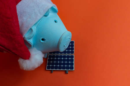 Piggy bank coins with Santa Claus christmas hat, photovoltaic solar panel and lamp isolated on orange background.の写真素材