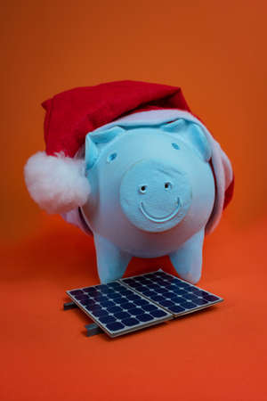 Piggy bank coins with Santa Claus christmas hat, photovoltaic solar panel and lamp isolated on orange background.の写真素材