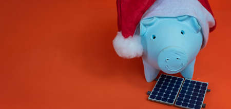 Piggy bank coins with Santa Claus christmas hat, photovoltaic solar panel and lamp isolated on orange background.の写真素材