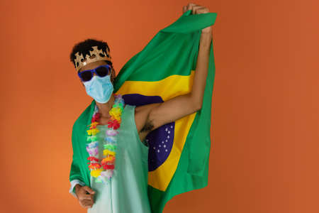 Black man in costume for carnival with brazil flag and pandemic mask isolated on orange background.の写真素材