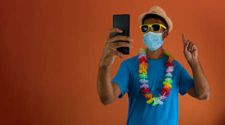 Black man in carnival costume and pandemic mask holding  a mobile phone isolated on orange background.の写真素材