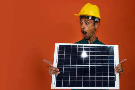 Worker With Helmet and Individual Protection Equipment Holding a Solar Photovoltaic Panel isolated. Solar Worker isolated in orange background.の写真素材