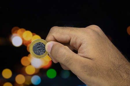 Holding a Brazilian money coin at a blurry lights background