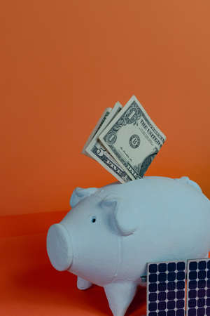 Piggy bank with dollar bill and photovoltaic solar panel isolated on orange background. Solar Energy Concept Image.の写真素材