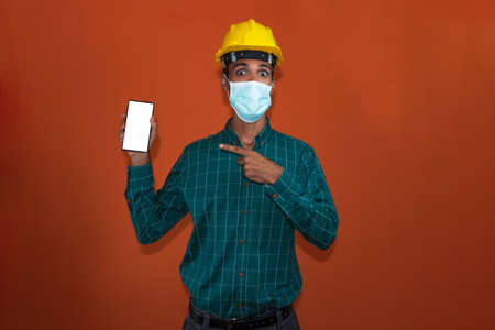 Engineer, architect or worker with yellow helmet and pandemic mask isolated on orange background. Engeneer at work holding a cell phone.の写真素材