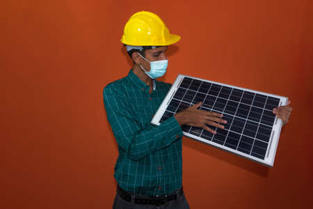 Engineer, architect or worker with yellow helmet and pandemic mask isolated on orange background. Engeneer at work holding a photovoltaic solar panel.の写真素材
