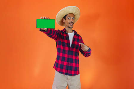 Black Man With Junina Party Outfit holding a tablet or mobile Isolated on Orange Background. Young...