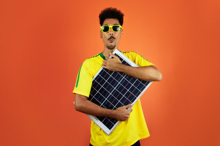 Soccer Player - Black Man Celebrating Holding Solar Photovoltaic Panel Isolated on Orangeの写真素材