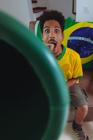 Black Man With Yellow Shirt in the Living Room watching TV cheering for Brazil. Happy Brazilian Man Watching Cup Game.の写真素材