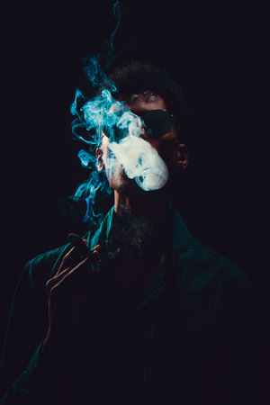 Black Young Man with Glasses Smoking Cigar with Fog at Black Background, low key light.