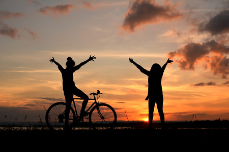 Silhouette of happy women with bicycle open hands in nature at the sky sunset color warm toneの写真素材