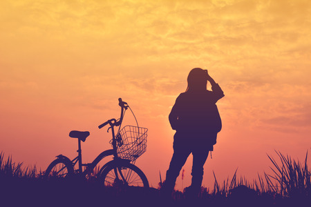 Silhouette of girl with bicycle on grass field at the sky sunset, color of vintage tone and soft focus concept journeyの写真素材