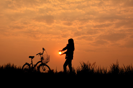 Silhouette of girl with bicycle on grass field at the sky sunset, color of vintage tone and soft focus concept journeyの写真素材