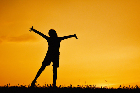 Silhouette of happy girl standing on grass field and sky sunsetの写真素材