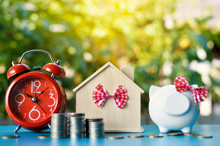 White piggy bank and alarm clock with wood house on table tree bokeh background ,concept save money ,selective and soft focusの写真素材