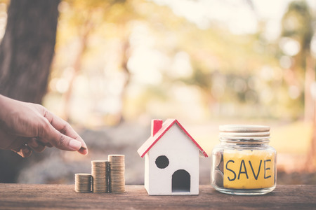Female hand holding coins on glass jar with save word and model house on table in nature bokeh...
