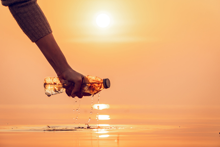Silhouette of human hand picking up bottle plastic in the lake outdoor on the baking hot day with sunrise morning, Drought and environmental problems and pollution concept.の写真素材
