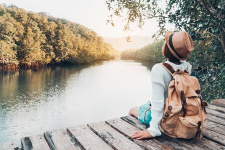 Cute girl backpacking and sitting on the wood plank resting and looking on the river.の写真素材