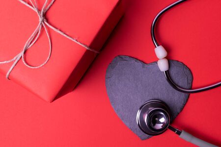 Heart of blackboard with stethoscope next to red gift box, on isolated red background.Concept give away health, world health day, take care of your heart.の写真素材