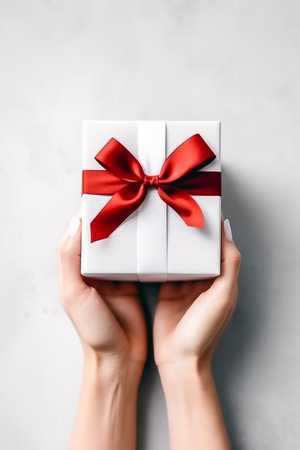 Female hands holding white gift box with red ribbon on gray background.の素材