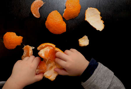 Children's hands are brushing mandarin on a black background. The child reaches for a slice of...