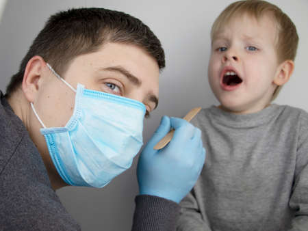 An otolaryngologist examines a child's throat with a wooden spatula. A possible diagnosis is inflammation of the pharynx, tonsils or pharyngitis. The concept of treatment and prevention of childhood diseasesの写真素材