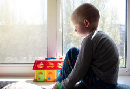 Pandemic isolation - outbreaks of coronovirus, covid-19. The child sadly looks out the window, sitting on a windowsill during quarantine. Quarantined children, home schooling, stay at homeの写真素材