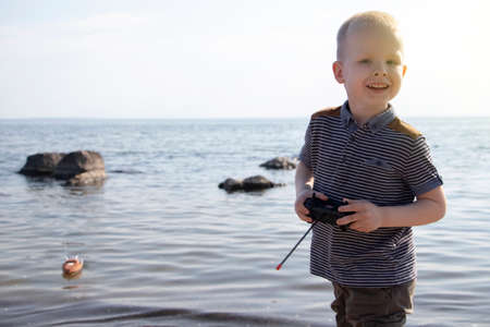 A blond-haired boy launches a radio-controlled ship. Game on the beach with a boat on the remote control. The concept of a holiday at sea and a happy childhoodの写真素材