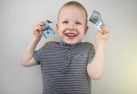 A blond boy holds dollars in his hands. The child rejoices in the appearance of pocket money. Financial literacy training conceptの写真素材