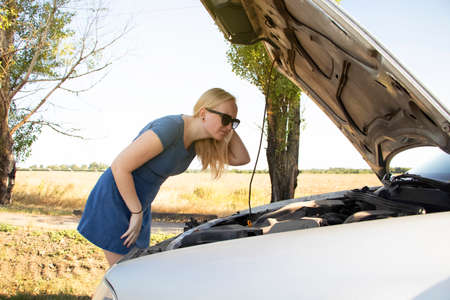 A blonde in a blue sundress votes on the road, near her car. An open bonnet indicates a breakdown in the vehicle. The concept of helping motorists in a difficult situation. Sweet girl in troubleの写真素材
