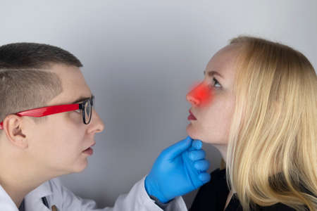 A young woman touches her nose, which is very painful. Medical care concept for difficulty breathing, clogged nasal passages and flu, colds or coronavirus. On examination by a doctor.の写真素材