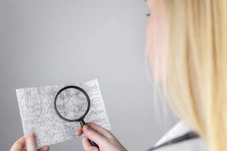 A neurologist with a magnifying glass examines an encephalogram of a patient's brain. Schedule of electroencephalograms - study of brain currents for signs of epilepsy and pathologiesの写真素材