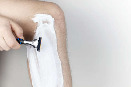 A man on a white background shaves his legs. Hairy legs and care for them. Gender equality concept. A close-up showing the foam, razor, and shaved hair on a smooth leg.の写真素材