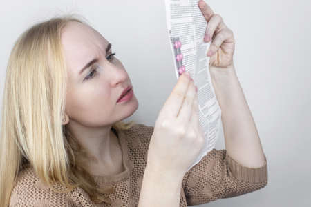 The girl on a white background reads the instructions for medicines. The woman looks at the list and composition of the drug. The concept of home self-medication and the study of the properties of drugsの写真素材