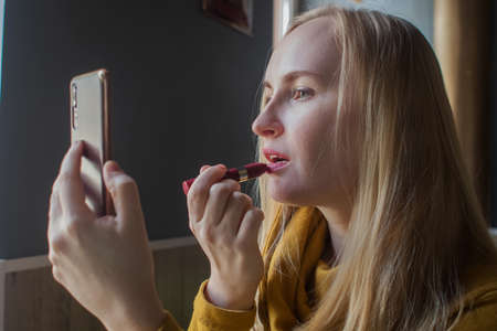The girl paints her lips in a cafe. Close-up of a cute girl who fixes her makeup after eating. The woman holds a mirror in a multi-colored frame. Personal care and make-up conceptの写真素材