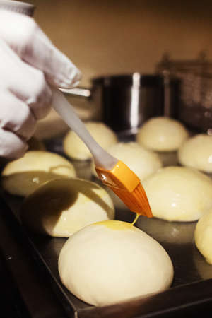 The baker greases the buns before placing them in the oven. Home baking and confectionery production. Home cooking concept. Vertical photoの写真素材