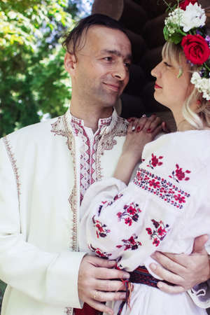 Young people are posing in traditional folk costumes. Cossacks man and woman in wreaths and embroidered shirts. Reconstruction of vintage traditions and customs. Guy and girl kissの写真素材