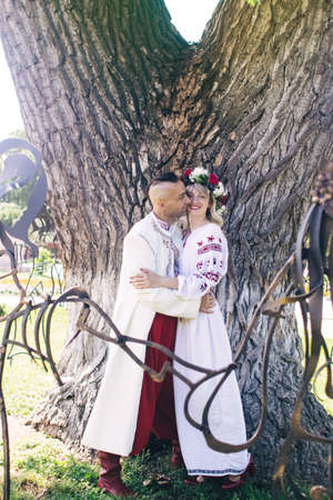 Young people are posing in traditional folk costumes. Cossacks man and woman in wreaths and embroidered shirts. Reconstruction of vintage traditions and customs. Guy and girl kissの写真素材
