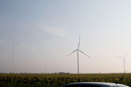 Windmills in the fog near a field of sunflowers. Early morning. See the movement of the blades, as a strong wind is blowing. Wind turbine are installed at different distances and hidden in the fogの写真素材