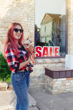 Close-up of a girl in fashionable clothes, who is considering purchases near the inscription SALE. Woman is holding a dog in a dress. Mini chihuahua. Shopaholism, shopping and glamorous life conceptの写真素材