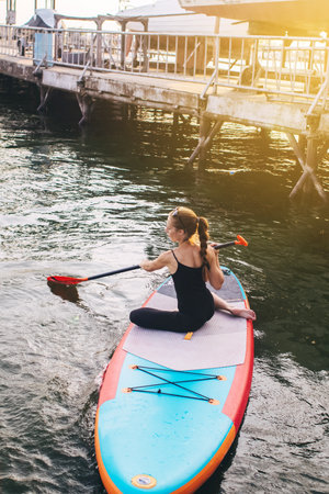Close-up of a girl doing yoga on sub board. Psychology meditation, relaxation and self-healing concept. Lonely woman alone on board against a background of water and a pier. Stand up paddle boardingの写真素材